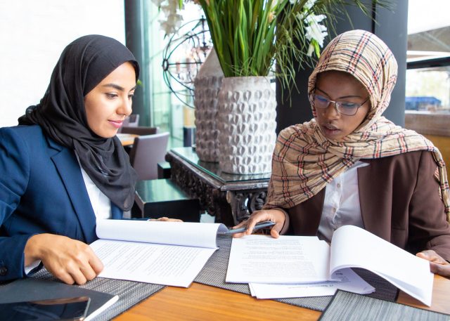 Confident female professionals checking documents. Young Muslim business women sitting in cafe and reading papers. Contract expertise concept